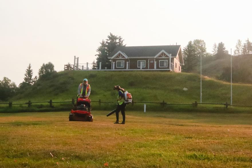 image of people maintaining the grass in an open field with a house in the background 