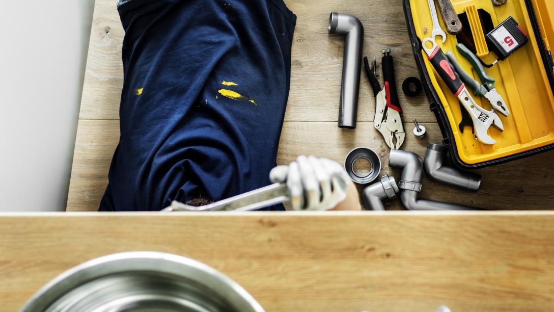 person under sink with plumbing fixtures and tools beside them in a yellow toolbox
