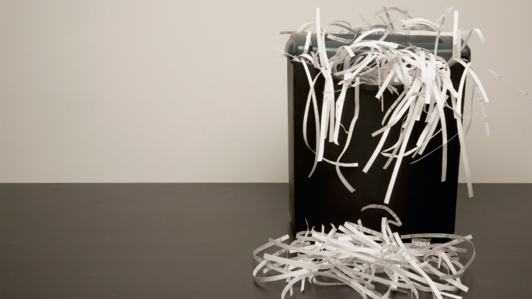 A black paper shredder overflowing with shredded white paper, with strips scattered on the floor in front.