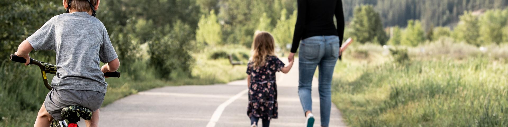 mom and daughter holding hands while son bikes behind them on pathway