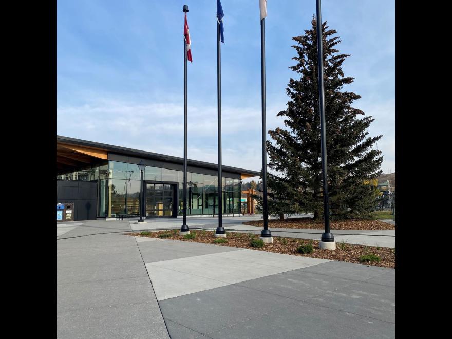 Row of three flagpoles outside of The Sation building. There is a tall Fir tree behind the flagpoles.