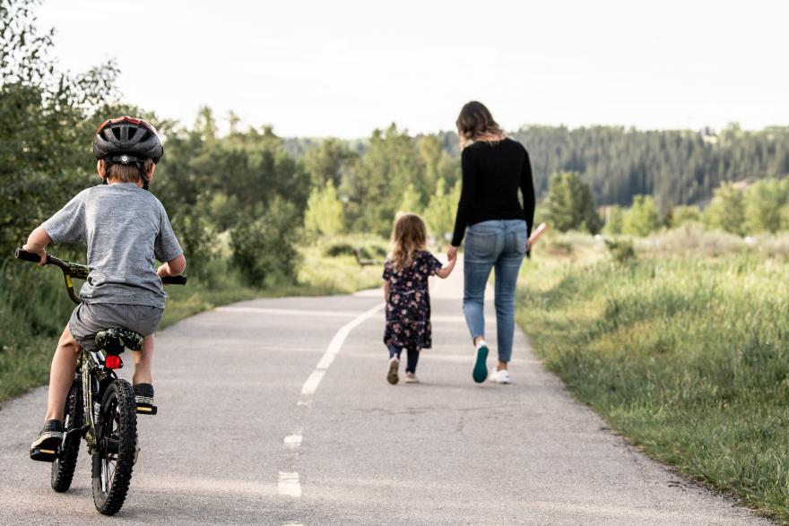 mom and daughter holding hands walking down pathway and son riding bike behind