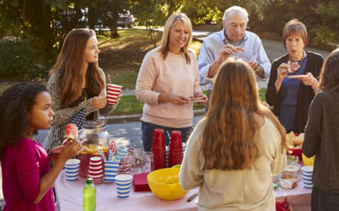 Group of people standing around a table with party favours. The people and table are outdoors near trees.