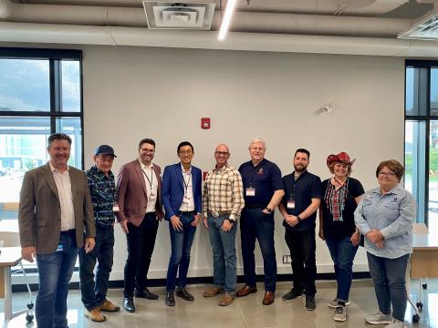 Group photo of men and women partaking in the Cochrane Business Incubator. The group of people are standing in front of a white wall and under an HVAC vent.