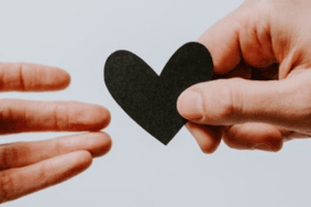 A hand handing out a black paper heart to another hand.