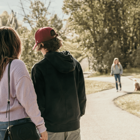 Two people standing in the forefront looking at something. They are giving their backs to the camera. In the background, there is a woman walking her dog on a paved pathway.