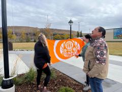 Two women holding up an orange truth and reconciliation flag with a man looking up at the flag pole.